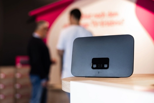 Ein WLAN-Router steht in einem modernen Telekom-Shop auf einem Tresen, zwei Personen im Hintergrund.